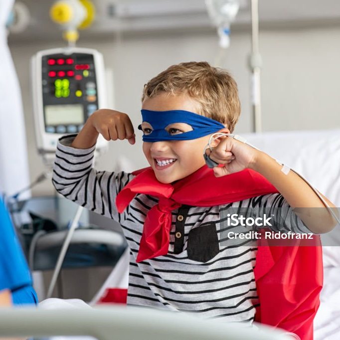 Cheerful strong little boy wearing blue eyeband and red cape like superhero sitting in hospital bed playing with nurse. Playful child gesturing dressed in superhero costume at clinic overcome adversity and health challenge. Happy smiling kid playing at hospital.