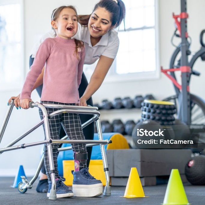 A little girl with a limb difference and a leg brace works with her physical therapist as she learns to use a walker.  The female therapist is bending down to aide the little girl as she directs her through a course.